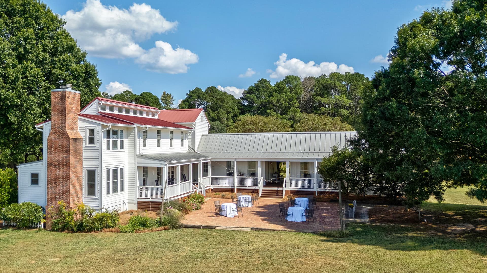 A large, white multi-story house with a red roof and a porch, surrounded by green trees and a grassy area, featuring several outdoor tables.