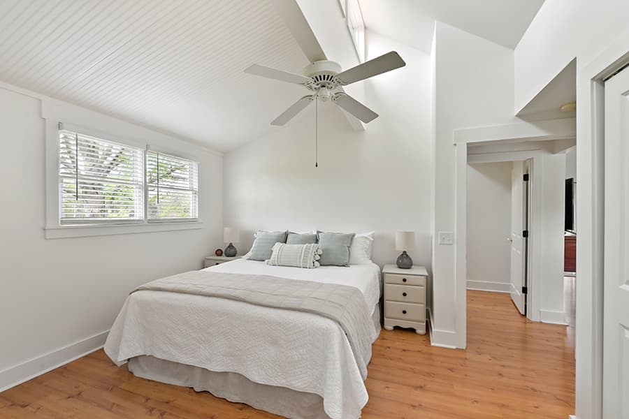 Bright, minimalist bedroom featuring a queen bed, ceiling fan, and natural light from windowed walls.