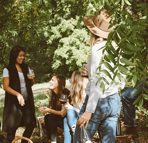 A group of four women laughs and enjoys drinks while sitting in a leafy outdoor setting.