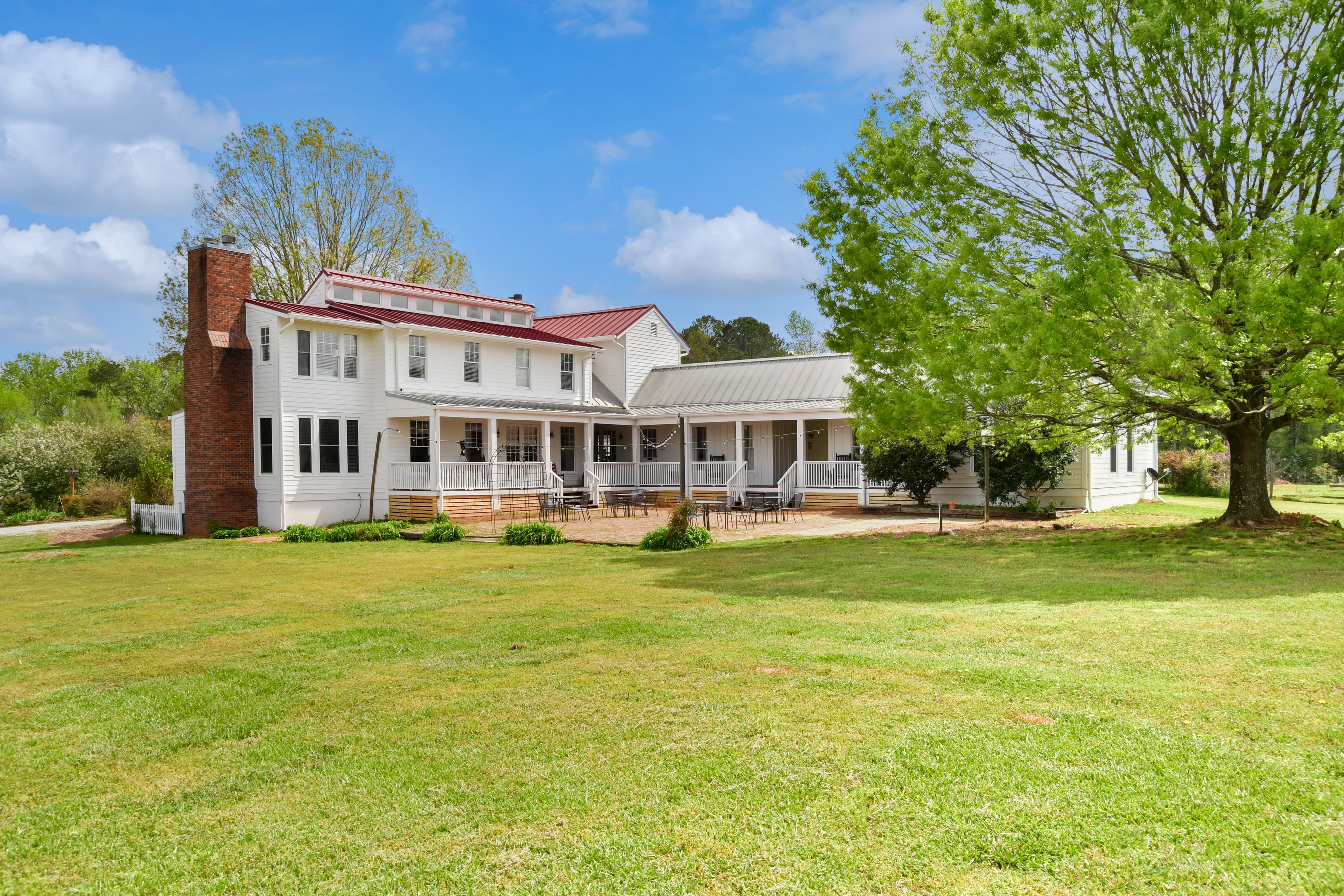 A spacious white house with a red roof set on a grassy lawn, surrounded by trees and blue skies.