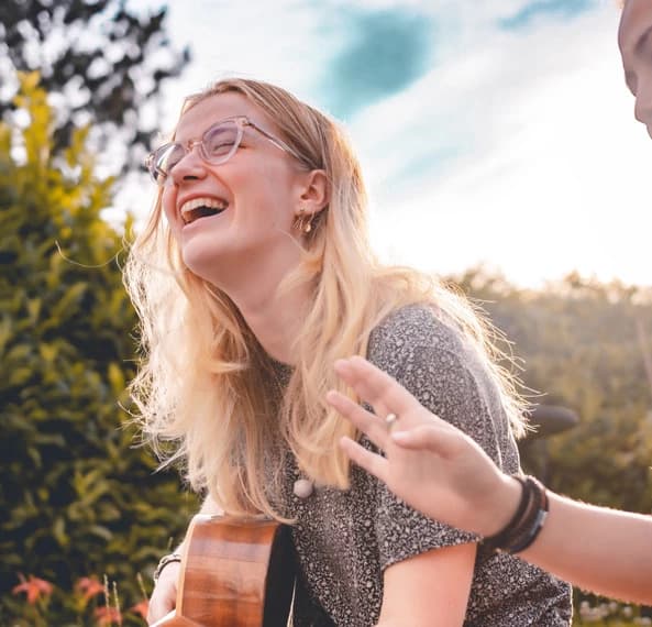 A smiling young woman with glasses laughs while holding a guitar outdoors.