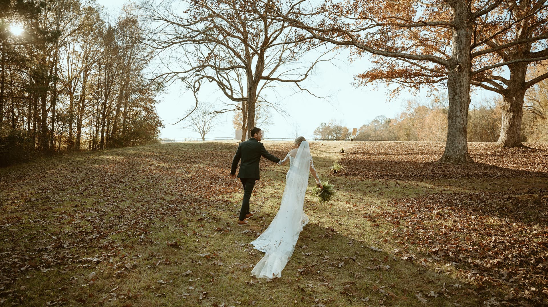 A couple walks hand in hand through a leaf-covered field, surrounded by trees.