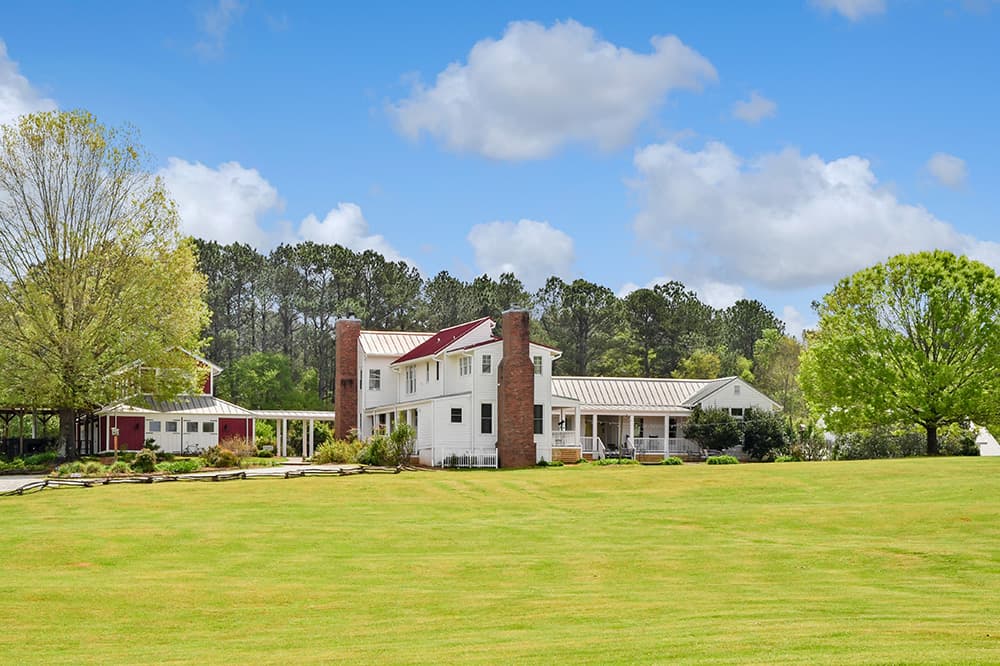 A large white farmhouse with red accents, surrounded by green fields and trees, under a blue sky.