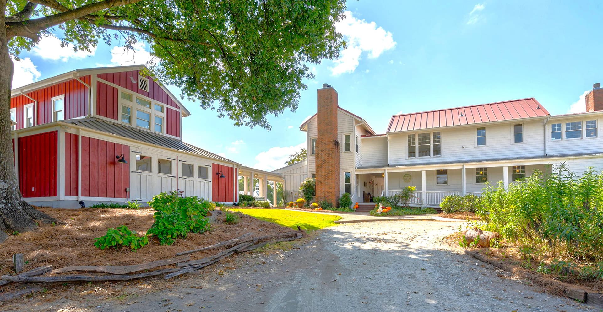 A scenic view of a rustic two-story home and a red barn-like structure, surrounded by green landscaping and a dirt driveway.