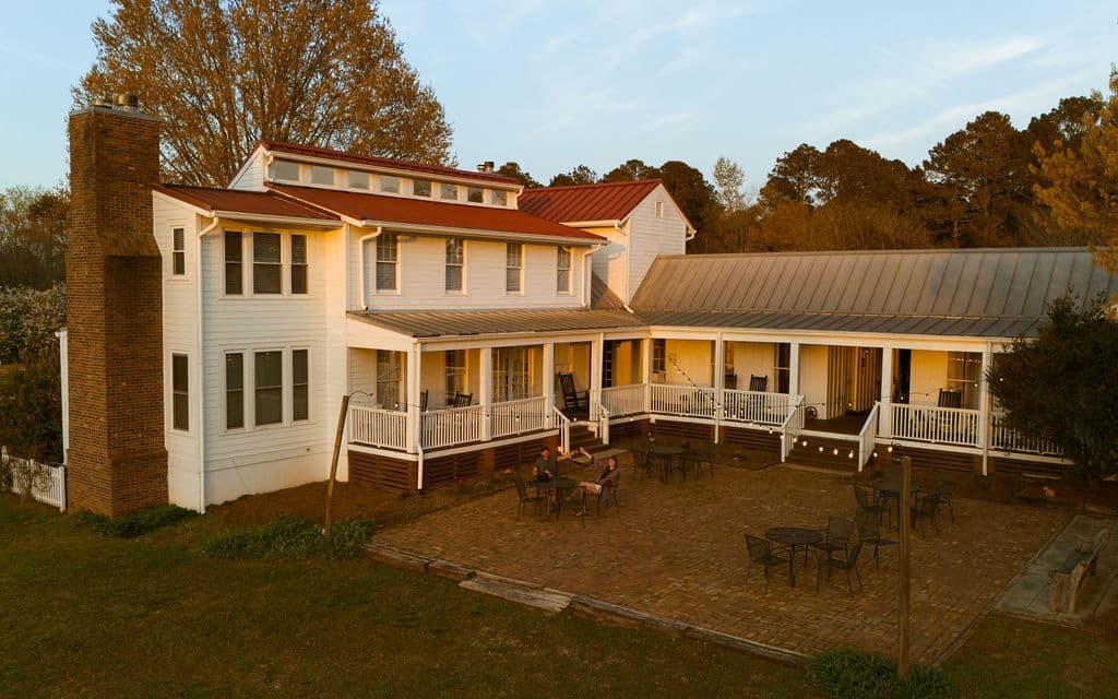A large, two-story white house with a red roof and a wraparound porch set in a grassy area.
