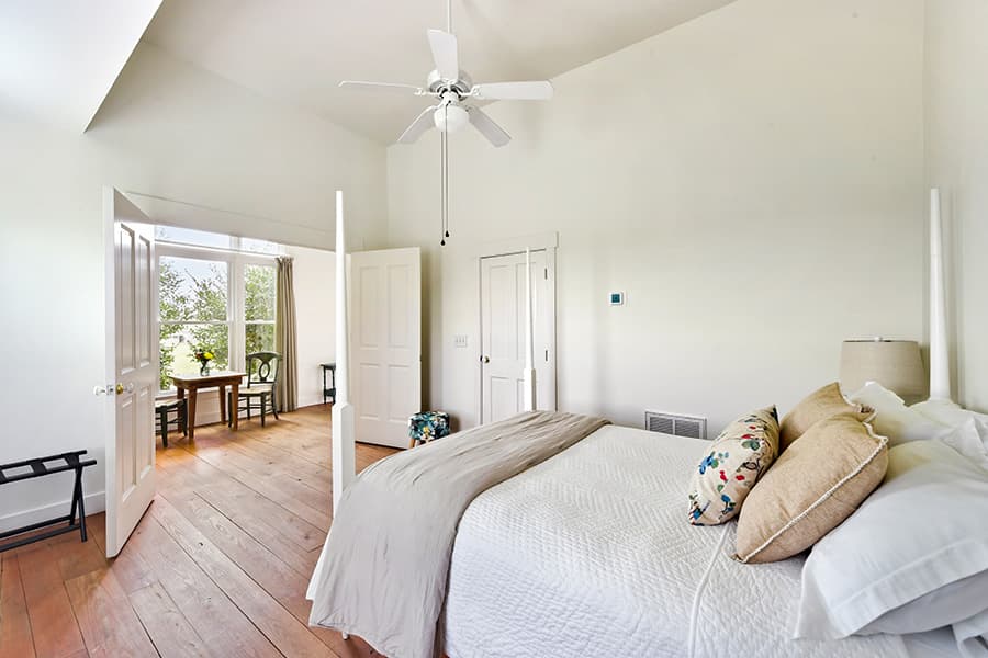 Bright, airy bedroom featuring a large bed, wooden flooring, and a view into a sunlit sitting area.