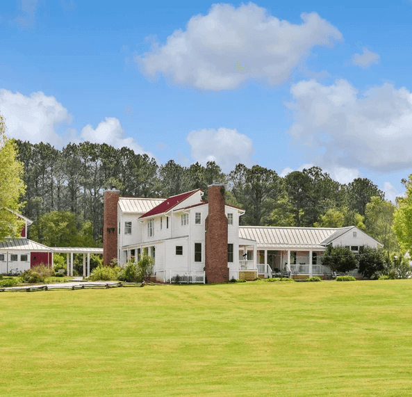 A spacious white farmhouse with a red roof surrounded by green grass and trees under a blue sky.